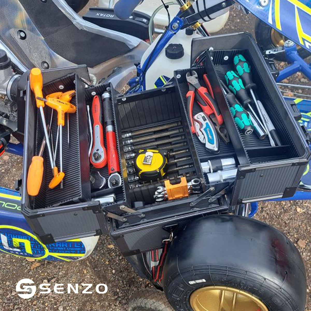 A black SENZO tool box with multiple compartments filled with various hand tools, shown mounted on a blue and yellow bicycle or scooter outdoors.